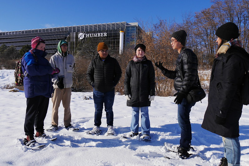 A group of students wearing snowshoes stand in a snowy field, the Humber Polytechnic visible on a building in the background.