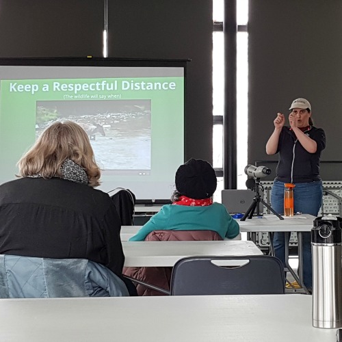A woman stands in front of a presentation screen that reads "Keep a Respectful Distance - The Wildlife Will Say When"