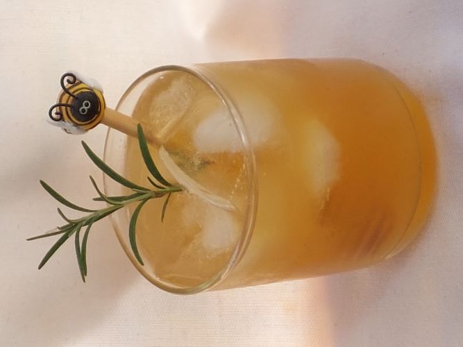 A glass with amber liquid, ice, a sprig of green, and a little swizzle stick topped with a bee