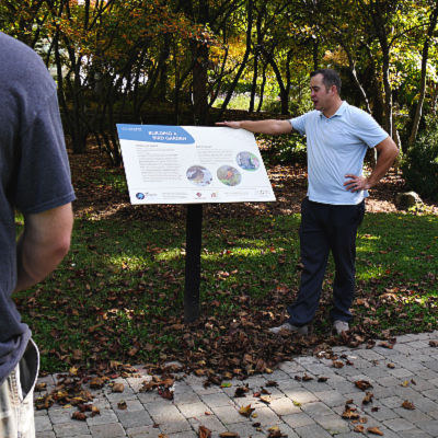 A rests his hand on an interpretive sign on the edge of a garden as he speaks to a group
