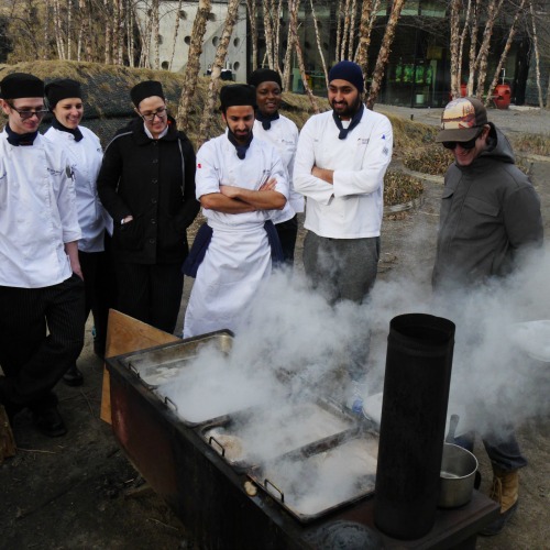 Students in chef's jackets and their instructor stand behind a steaming maple syrup evaporator with an educator from the Arboretum