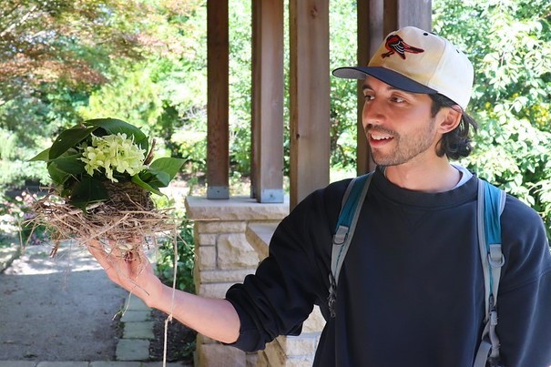 A man in on Orioles baseball hat smiles as he holds up and looks at an elaborate nest-like structure made of natural materials by children at a nature camp.