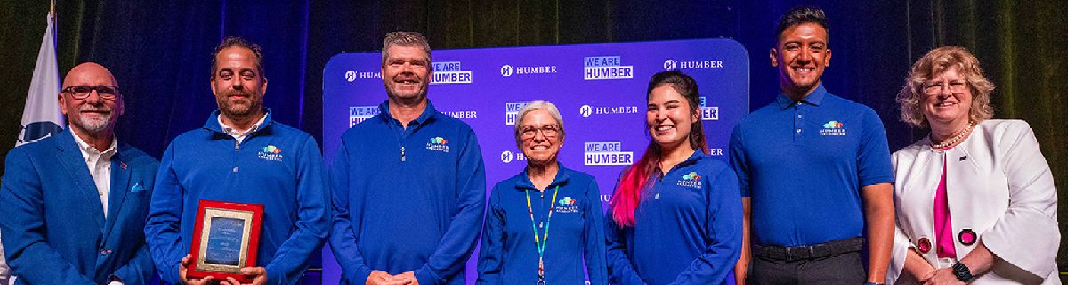 Five members of the Arboretum team in their staff shirts, one of whom is holding an award, stand on a stage flanked by two other people, all smiling for the camera