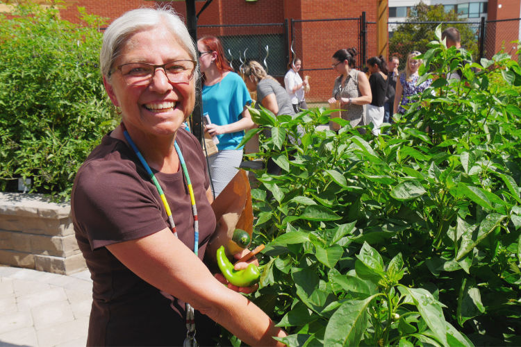 A woman holding a pepper in a large vegetable garden smiles at the camera while other people mill around in the background