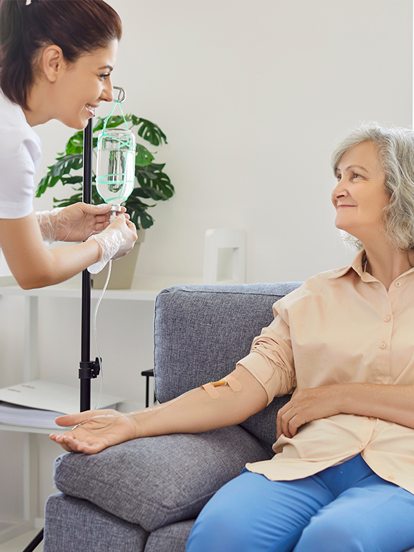 Senior woman with intravenous drip sitting on couch, physician, nurse doctor fixing IV drip infusion