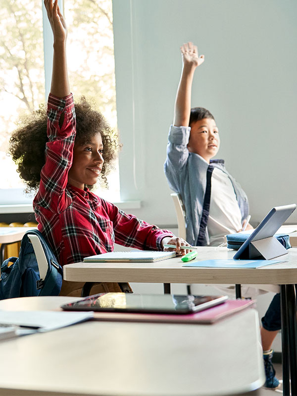 two students sitting a desks with there hands raised