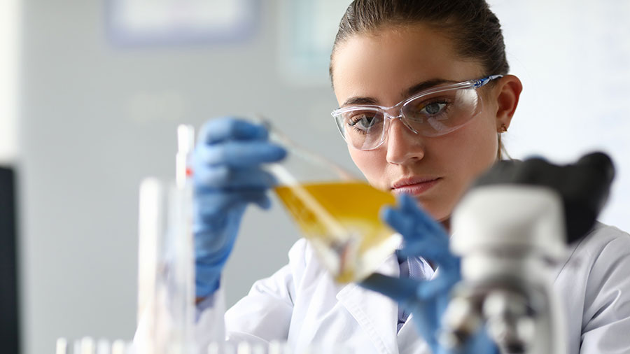 Close-up of laboratory researcher holding medical glass bottle with yellow liquid wearing sterile gloves and special lab eyewear