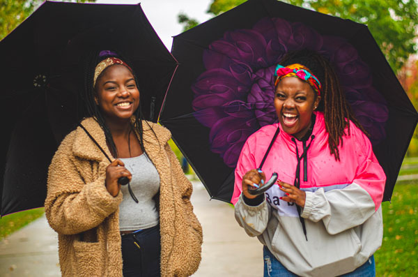 two students outdoors smiling and holding umbrellas