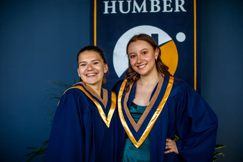 Two graduates pose for photo in front of Humber flag