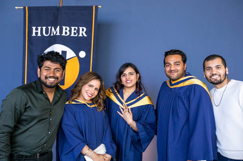 Three graduates and two guests take photo in front of Humber flag