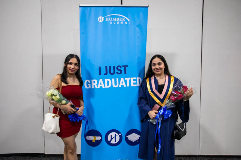 Two graduates holding flowers stand beside banner that reads I Just Graduated