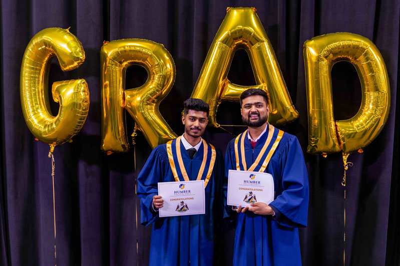 Student posing for picture with diplomas in front of Grad Balloons
