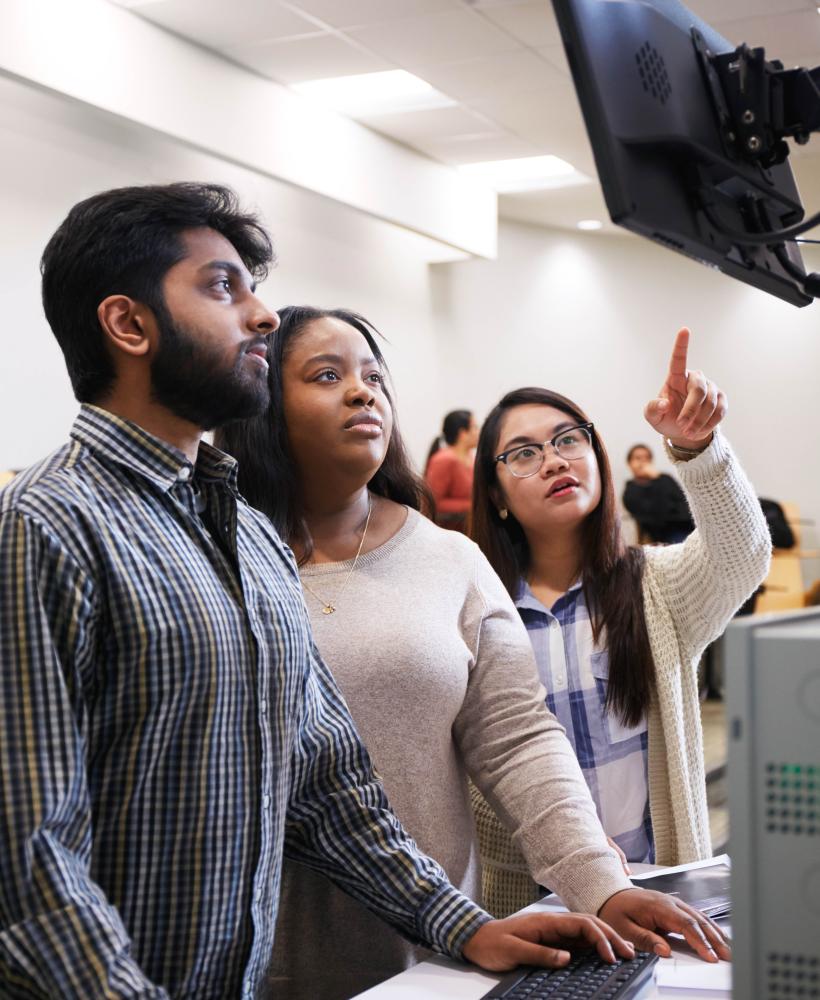Three students looking up a monitor mounted high on a wall - one student is pointing at something on the screen.