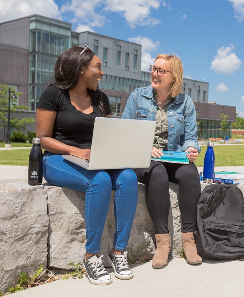 Two students sitting on a rock ledge at the Lakeshore Campus - one has a laptop on her lap