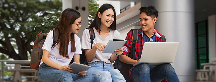 Image: Students sitting outdoors on the university campus, smiling and socializing, using their laptop and mobile devices.