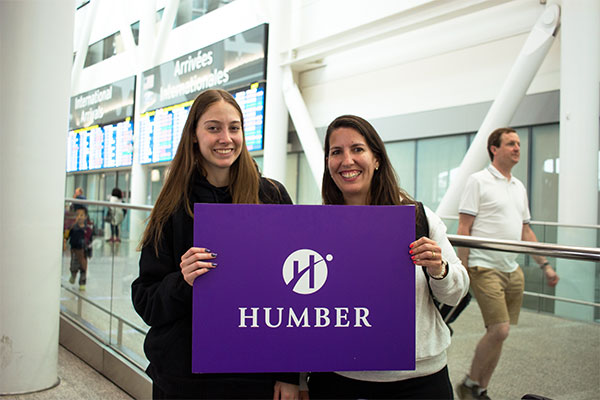2 students at airport holding Humber sign