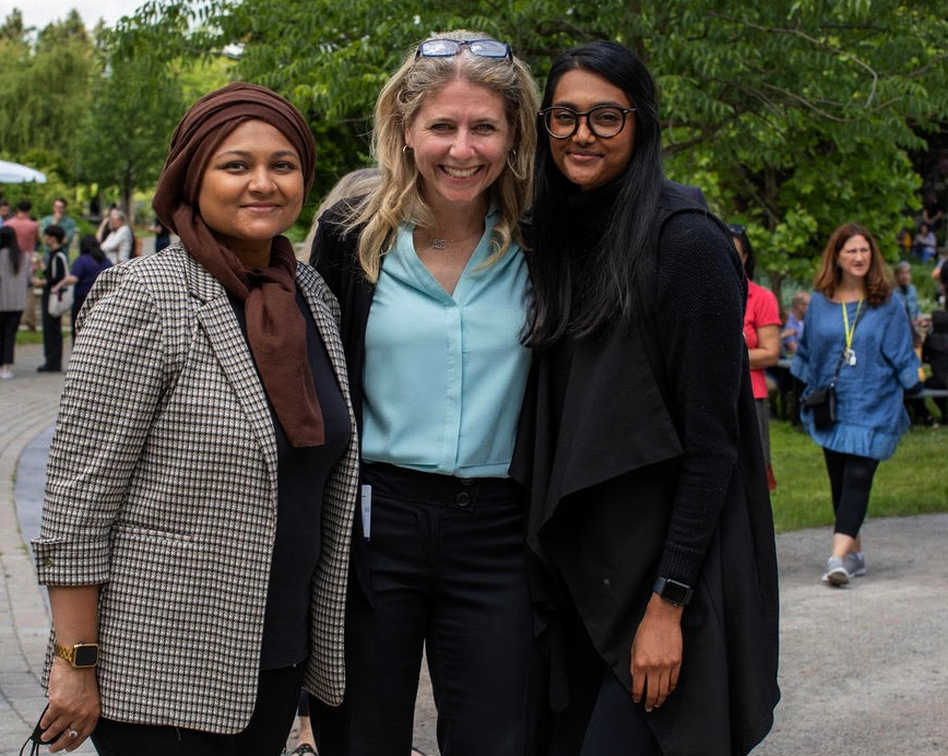 Three women standing close together on a path with trees in the background.