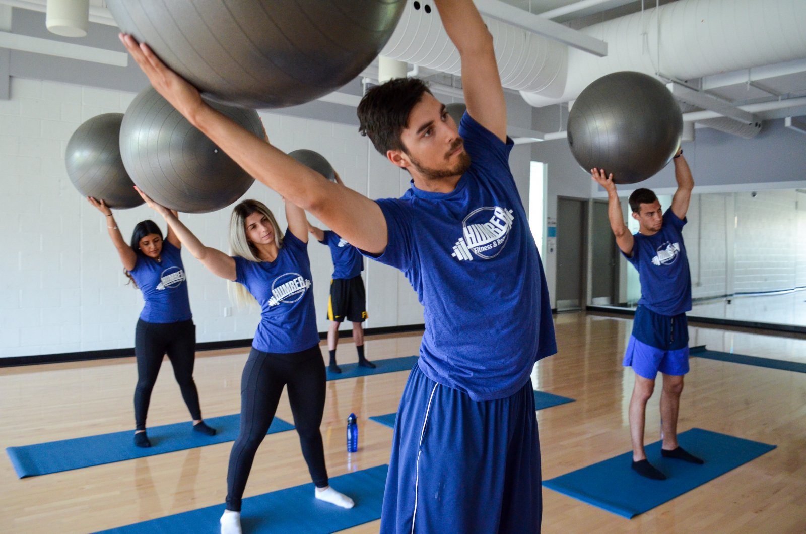 A gym glass - students dressed in blue shirts hold exercise balls over their heads.