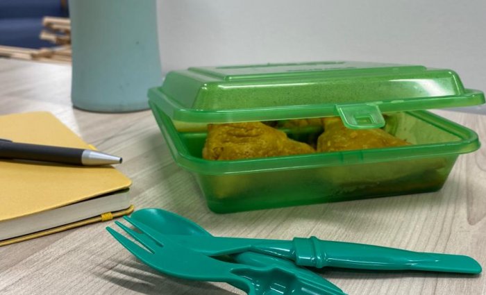 A reusable container with food inside it sits on a desk with a reusable water bottle and reusable cutlery near it.