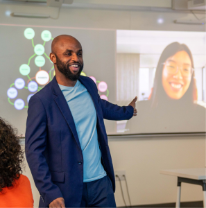 A man in a suit gestures towards a virtual participant displayed on a screen during a hybrid learning session in a classroom.