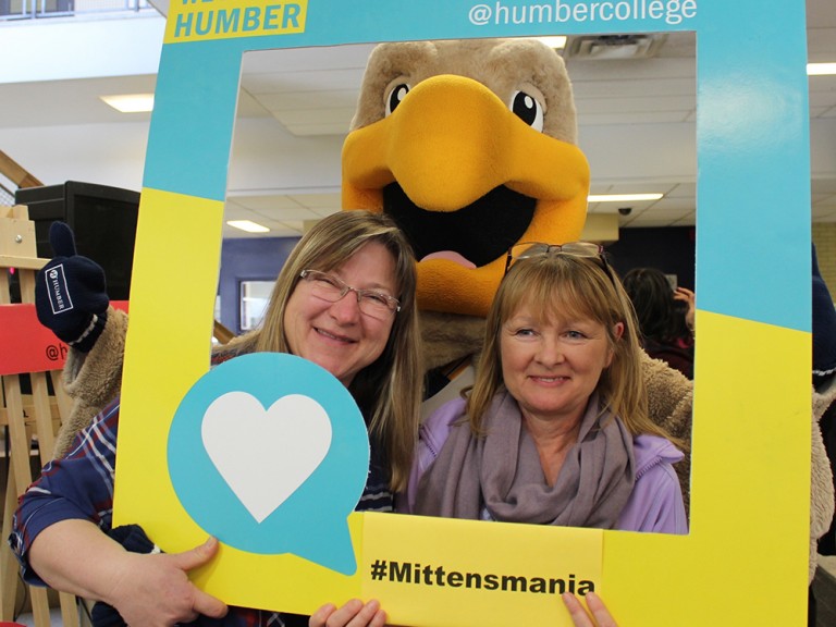 two women posing behind a photo frame with the humber hawk mascot
