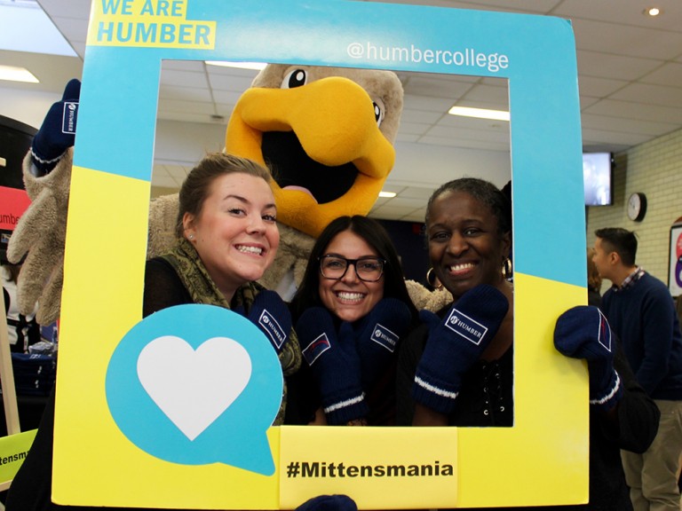 three women posing behind a photo frame with the humber hawk mascot