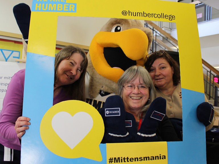 three women wearing mittens and posing with the humber hawk mascot behind a photo frame