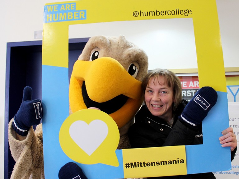 a woman wearing mittens posing behind the photo frame with the humber hawk mascot