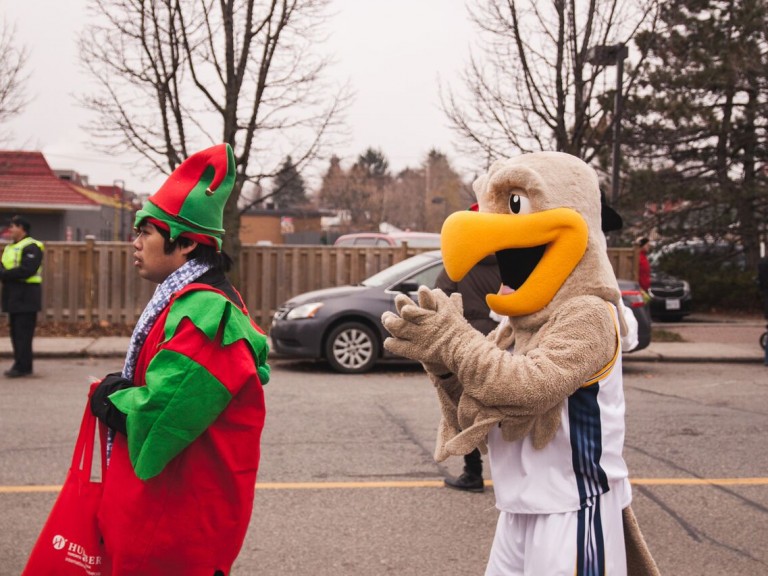 humber hawk mascot walking behind a person in an elf costume