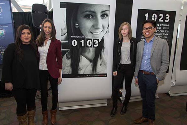 four people standing in front of a banner with a girl's face that says fentanyl death number 0103