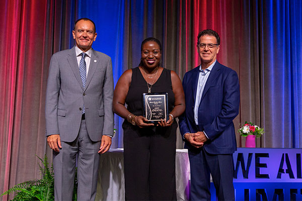 Award recipient from 2018 holding a plaque beside 2 presenters