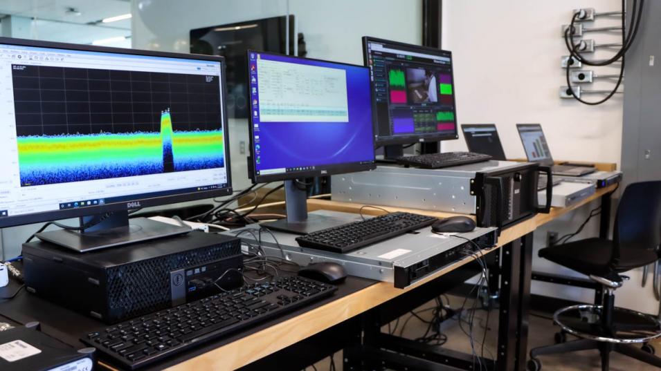 Three wide computer screens sit atop large computers on a long desk in the B2C lab. They show colourful graphs and and documents. There is a large window behind the computers showing a glimpse of the next room.