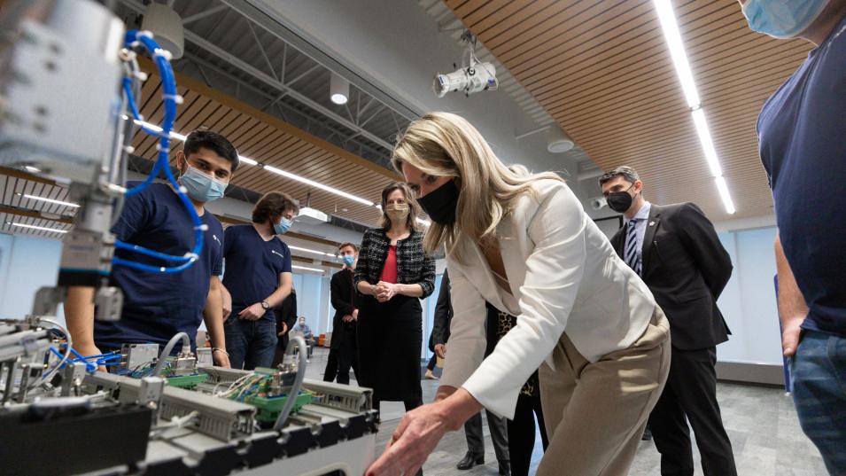 Masked MPP Jill Dunlop bends over a large, FESTO-branded machine with students and officials in the background, all masked