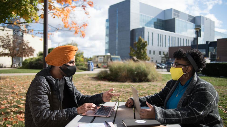 Two students sit across from each other at a picnic table on the lawn with Humber's LRC building in the background