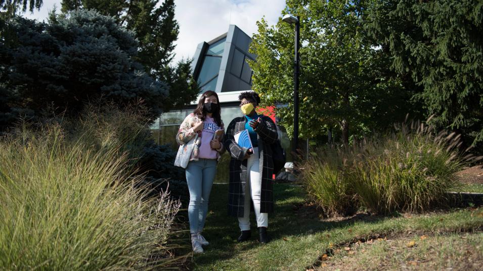 Two students wearing masks walk outside amidst grass and green foliage with the Humber Arboretum in the background