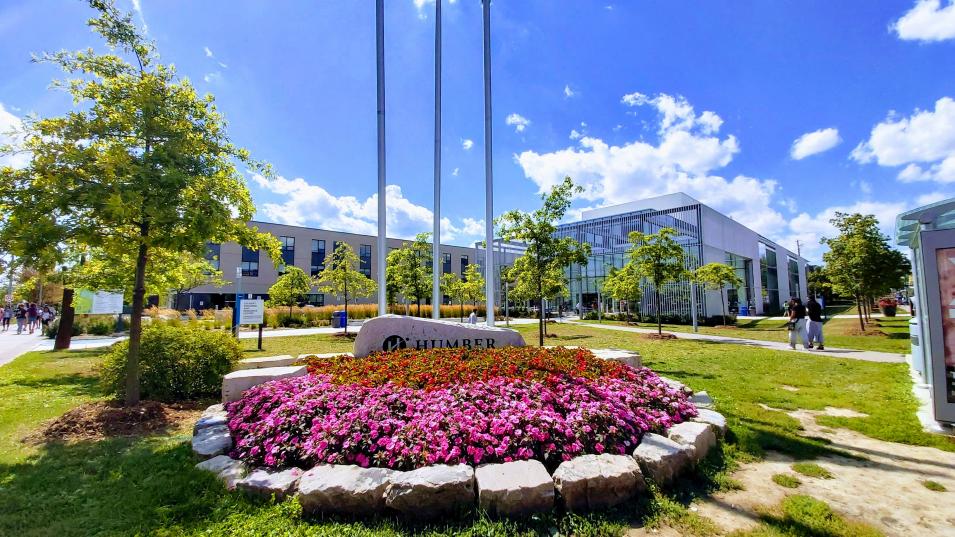 A photo by Mohammed Taabish shows a bed of flowers around a rock with 'Humber' written on it, in front of three flagpoles at the Humber Lakeshore campus entrance.