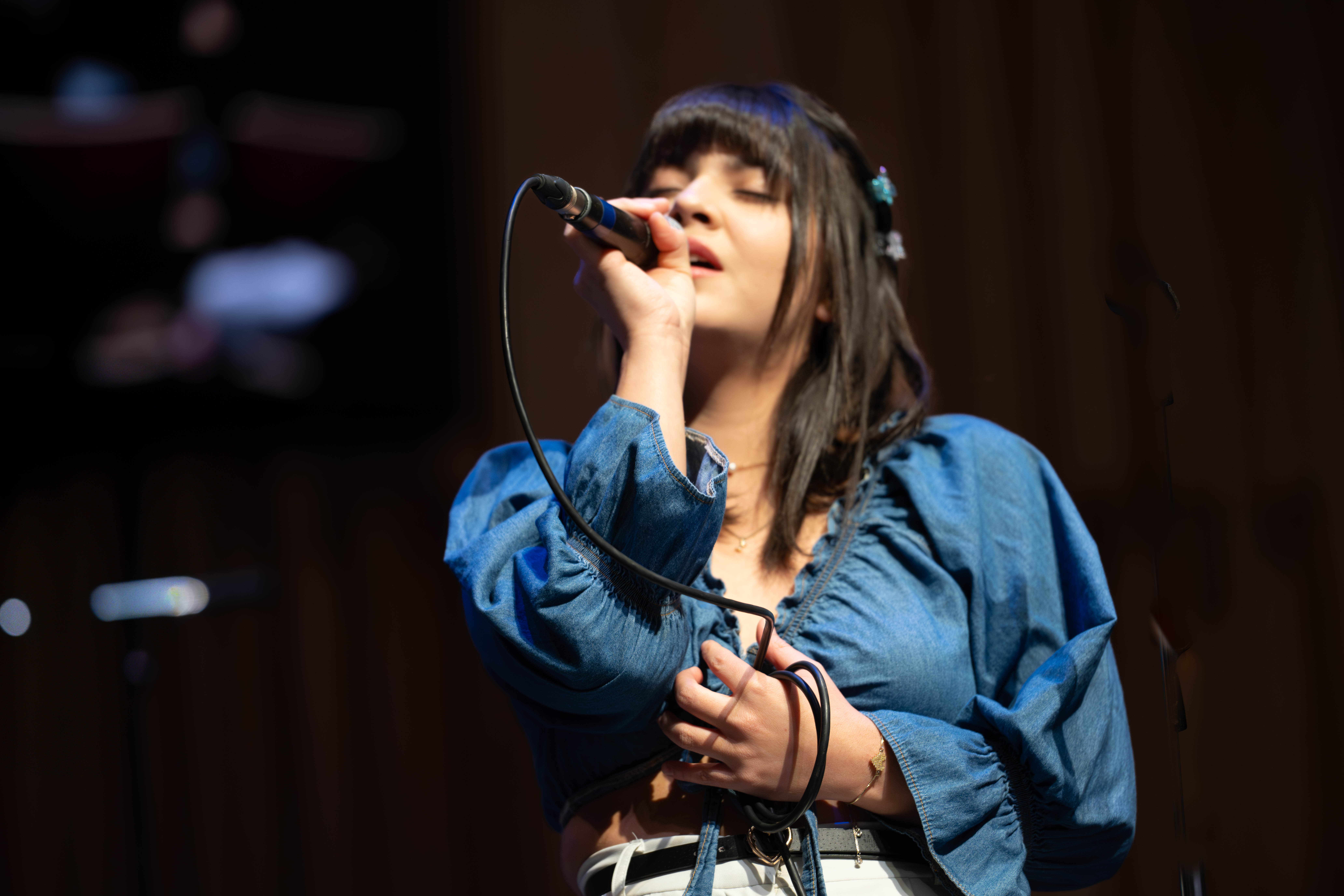 Young woman with black hair, denim blouse and white trousers sings into a microphone on stage
