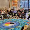A large group of people sit in a circle around a brightly coloured rug with flowers and other designs on it.