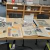 Bound copies of old newspapers sit on a desk. Photographs can be seen in the background on another table.