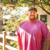 Scott Iles wears a pink shirt and poses in front of a fence and some foliage.