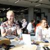 Smiling people sit at a table that’s covered in paper and other items.