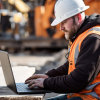 A person wearing a hardhat and reflective vest works on a laptop at a construction site.