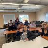 A group of people sit at desks while waving to the camera.