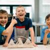 Three smiling youth are building a miniature bridge using sticks.