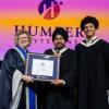 Three people stand together on a stage. Two are holding the same honorary degree.