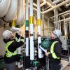 Two people wearing hard hats and high visibility vests are looking at a series of pipes.