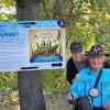 Two people are in a forest beside a sign that reads StoryWalk.