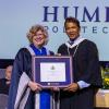 Two people wearing graduation robes stand together and are holding an honorary degree.