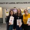 Three smiling people hold small posters while standing beneath a sign that reads Faculty of Liberals Arts & Sciences.