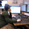 A person sits at a desk and looks at a computer screen. A person stands beside them while working.